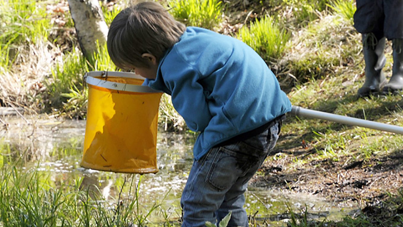 En pojke hovar vid ett vattendrag. En vuxen står en bit ifrån.